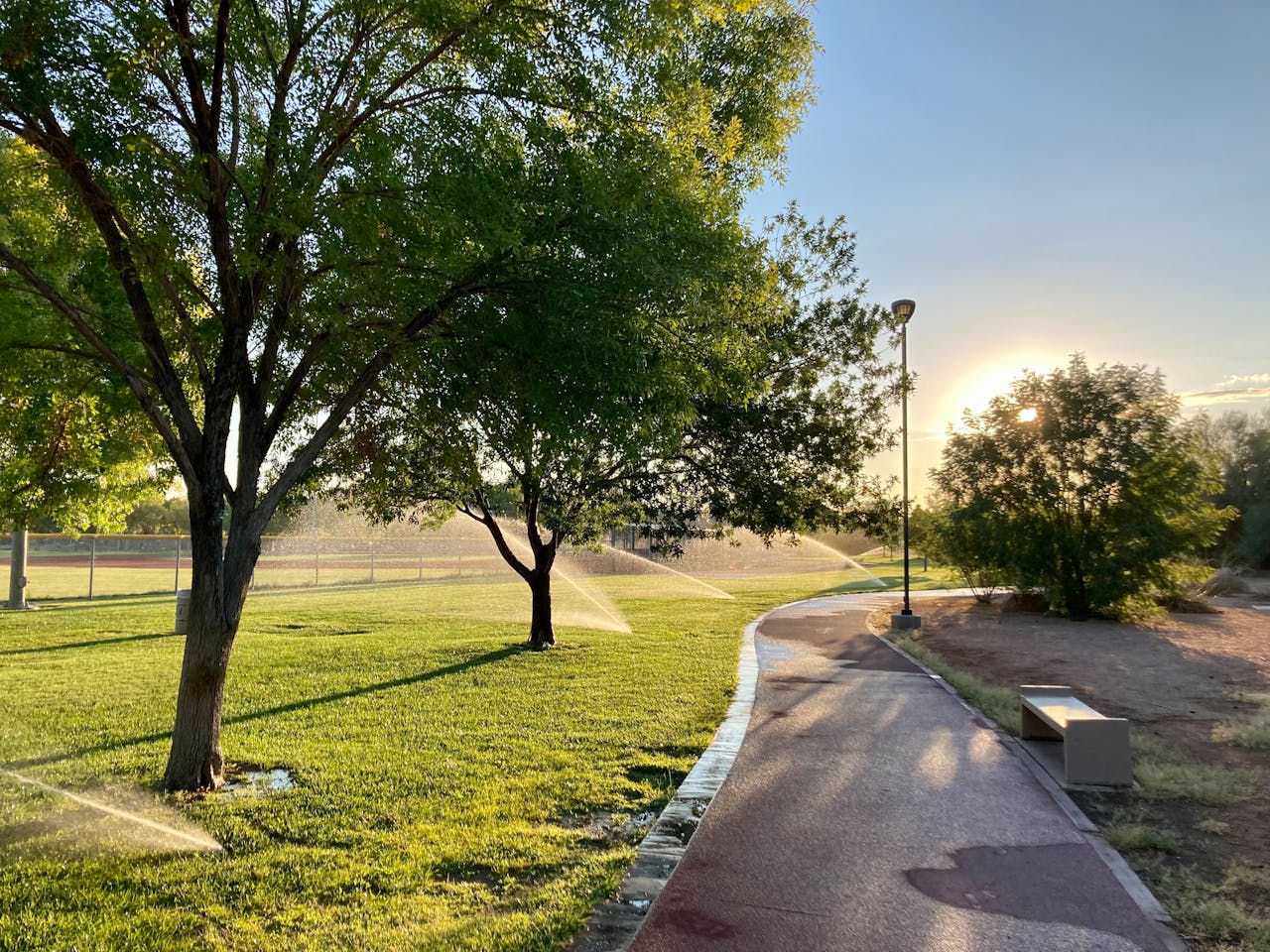 our-services-2 Sunlit park with pathway and sprinklers watering lush green grass and trees.